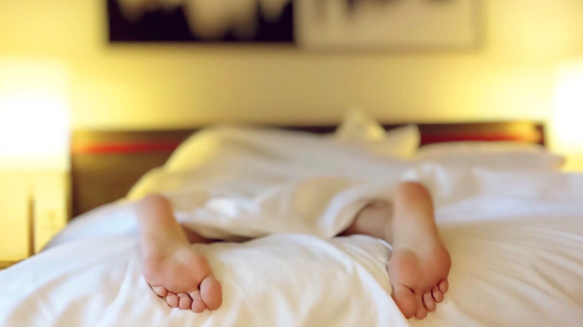 Person lying in bed face down under a white blanket with their feet sticking out.