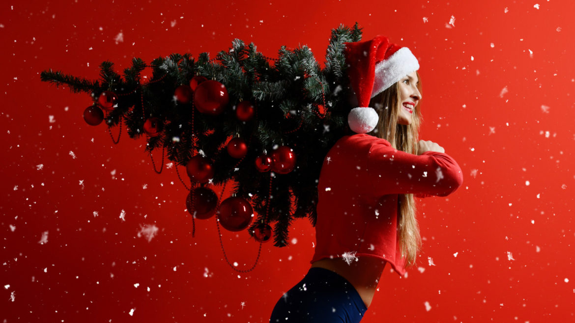 Woman wearing santa hat while carrying a christmas tree on her shoulders and snowflakes falling.