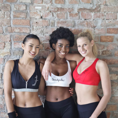 Three diverse women wearing sports bras and leggings while standing in front of a brick wall.