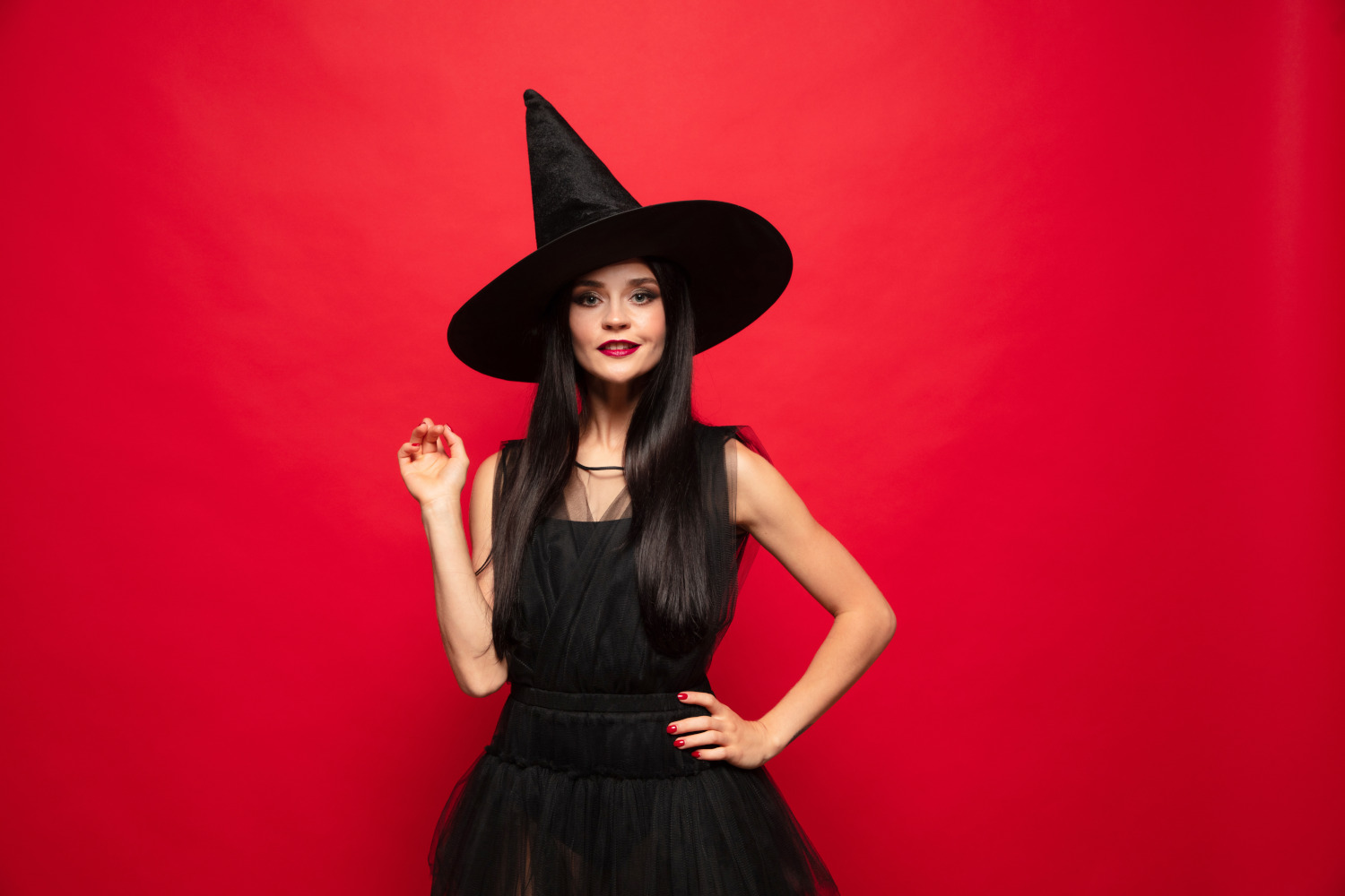 Brunette woman in witch costume with a black pointy on a red background pointing up.