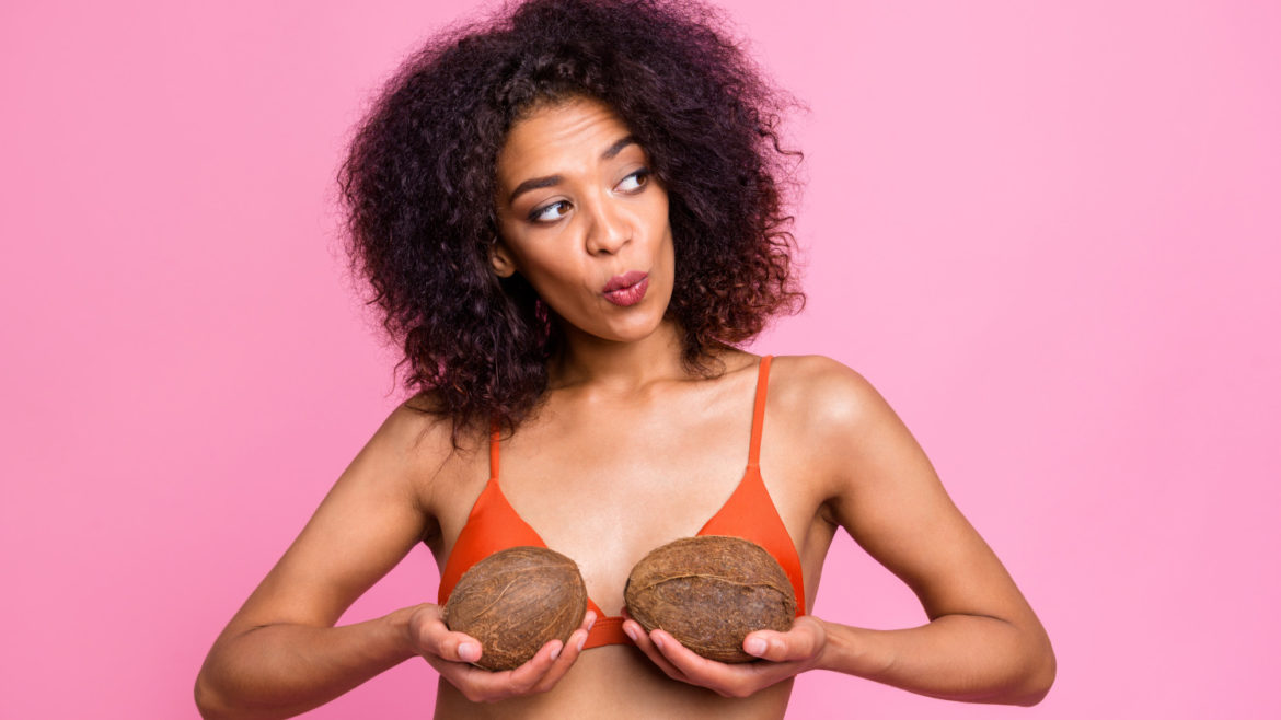 Trendy woman wearing orange bikini while holding coconuts up to try on new breast size in front of isolated pink background.