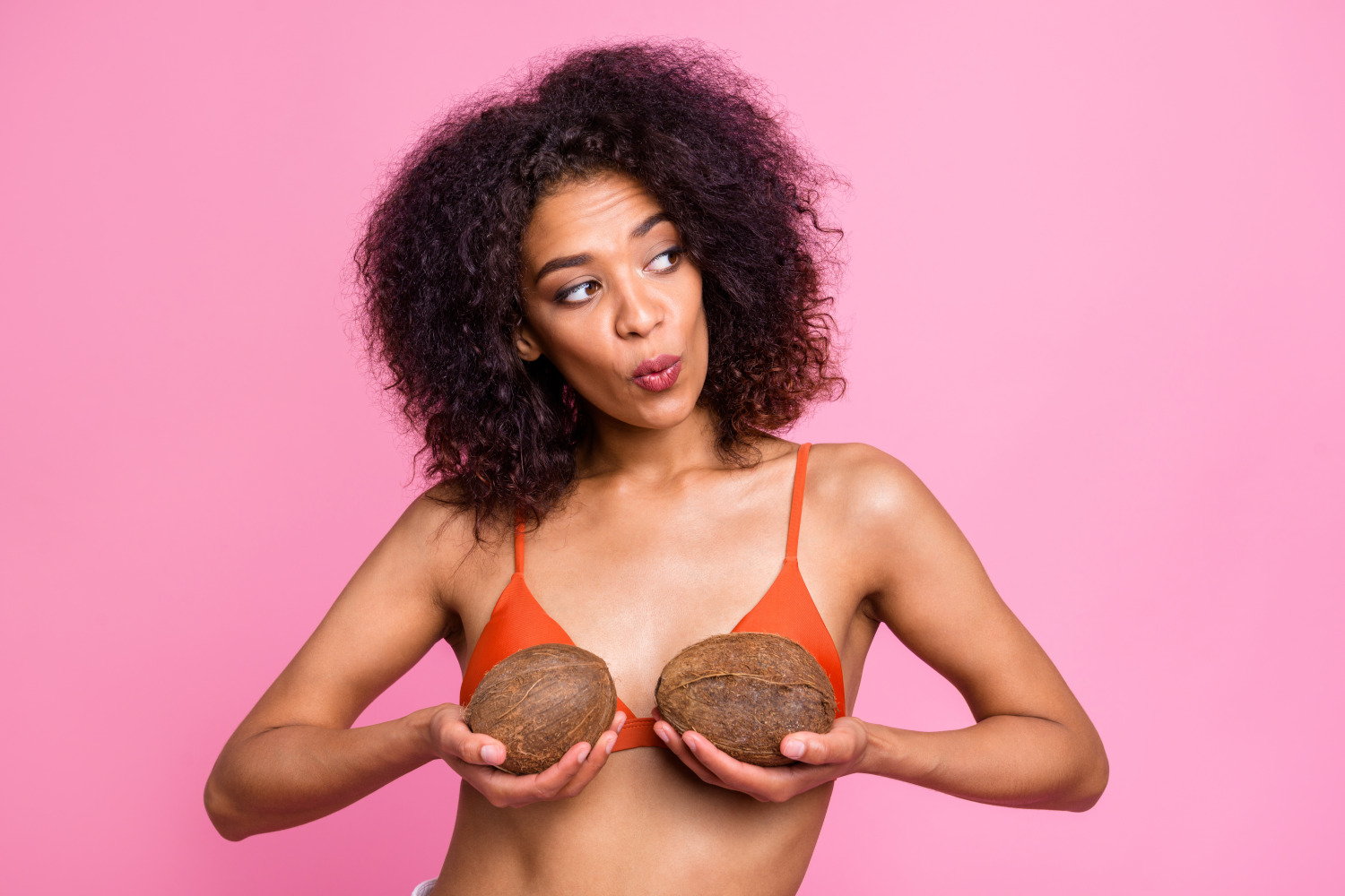 Trendy woman wearing orange bikini while holding coconuts up to try on new breast size in front of isolated pink background.