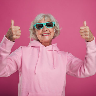 Positive old woman in pink hoody and black with green sunglasses posing with two thumbs up on pink background.