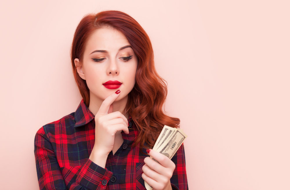 Surprised redhead girl with money in hand on a pink background.