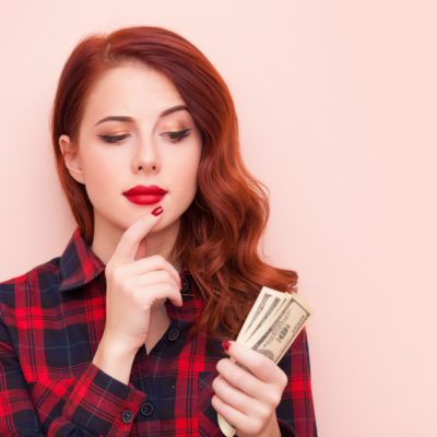 Surprised redhead girl with money in hand on a pink background.