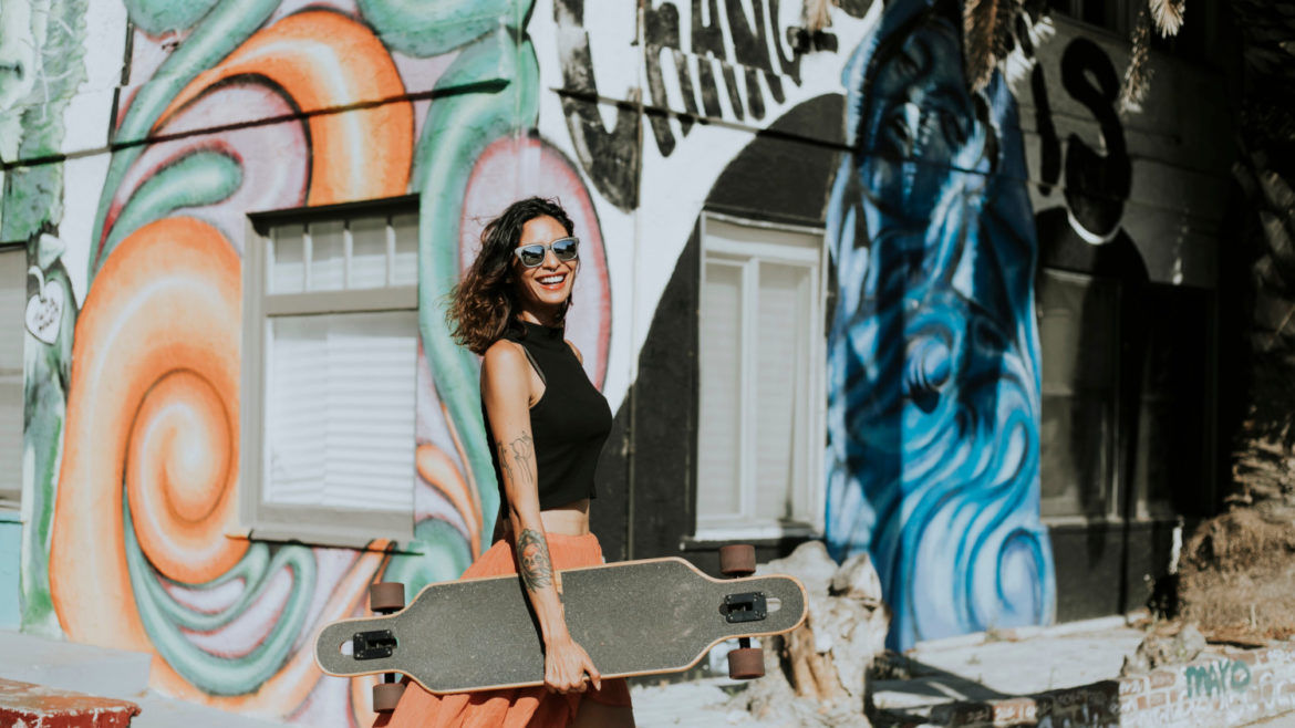 Woman in black crop top and orange flowing skirt holding a longboard walking throught the city.