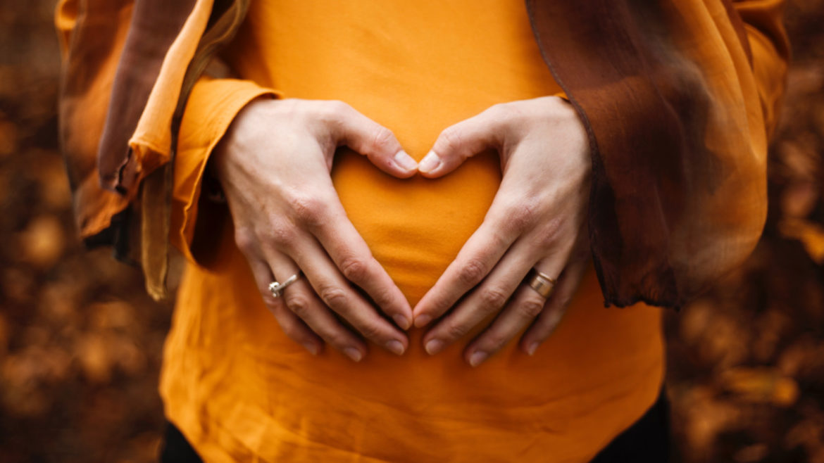 Woman in an orange colored blouse touching her pregnant tummy.