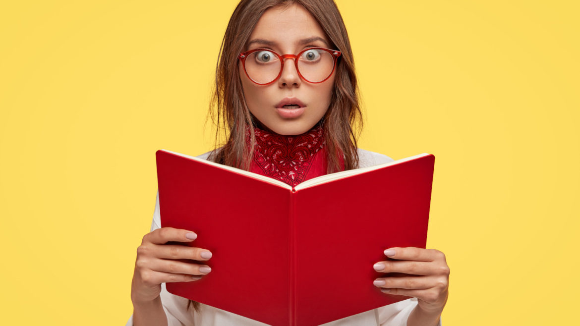 Lovely woman wearing glasses holding a book looking concerned about the information she is reading in front of an isolated yellow background.
