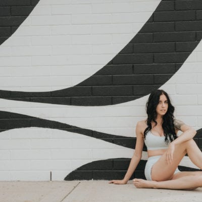 Woman in an white sports bra and shorts posing in front of a black and white swirled wall. Nonsurgical mommy makeover concept.