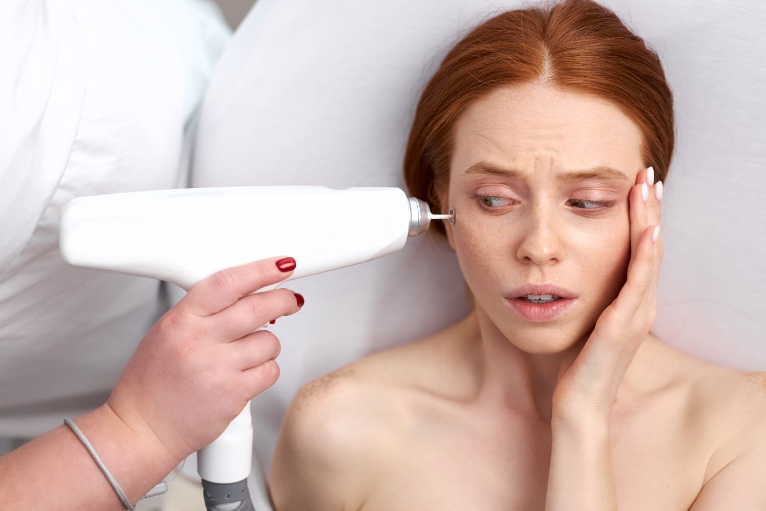 Red haired woman scared by laser treatment on face, looking at equipment.
