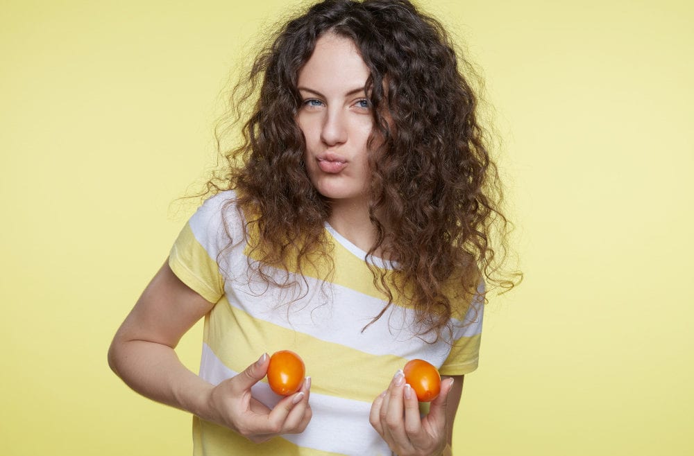 Young woman in yellow and white striped shirt holds tomatoes in hands above her breasts.